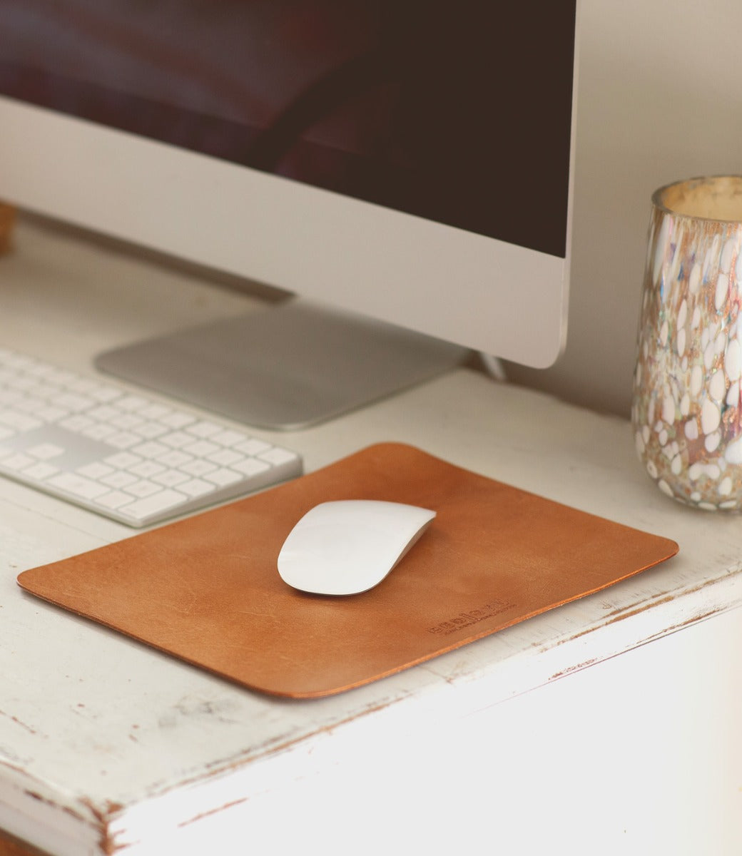A computer mouse rests on a brown leather **Bed Stu Launcher Mouse Pad**, boasting a suede underside, next to an Apple keyboard and monitor. A decorative cup is placed to the right of the setup on a white desk, completing the carefully curated workspace.