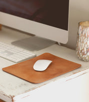 A computer mouse rests on a brown leather **Bed Stu Launcher Mouse Pad**, boasting a suede underside, next to an Apple keyboard and monitor. A decorative cup is placed to the right of the setup on a white desk, completing the carefully curated workspace.
