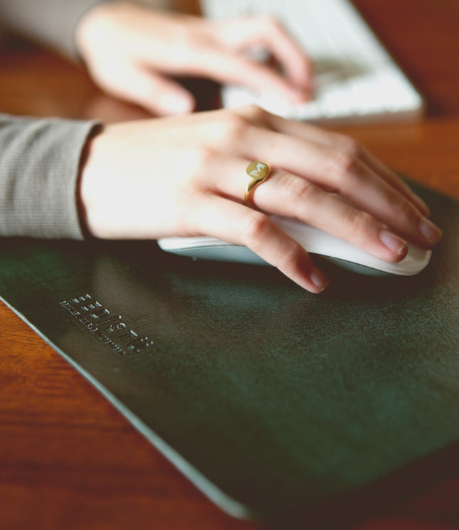 A person's hand, wearing a ring, rests on a computer mouse with a keyboard in the background. The mouse is on a Bed Stu Launcher Mouse Pad with a suede underside, enhancing the curated workspace.