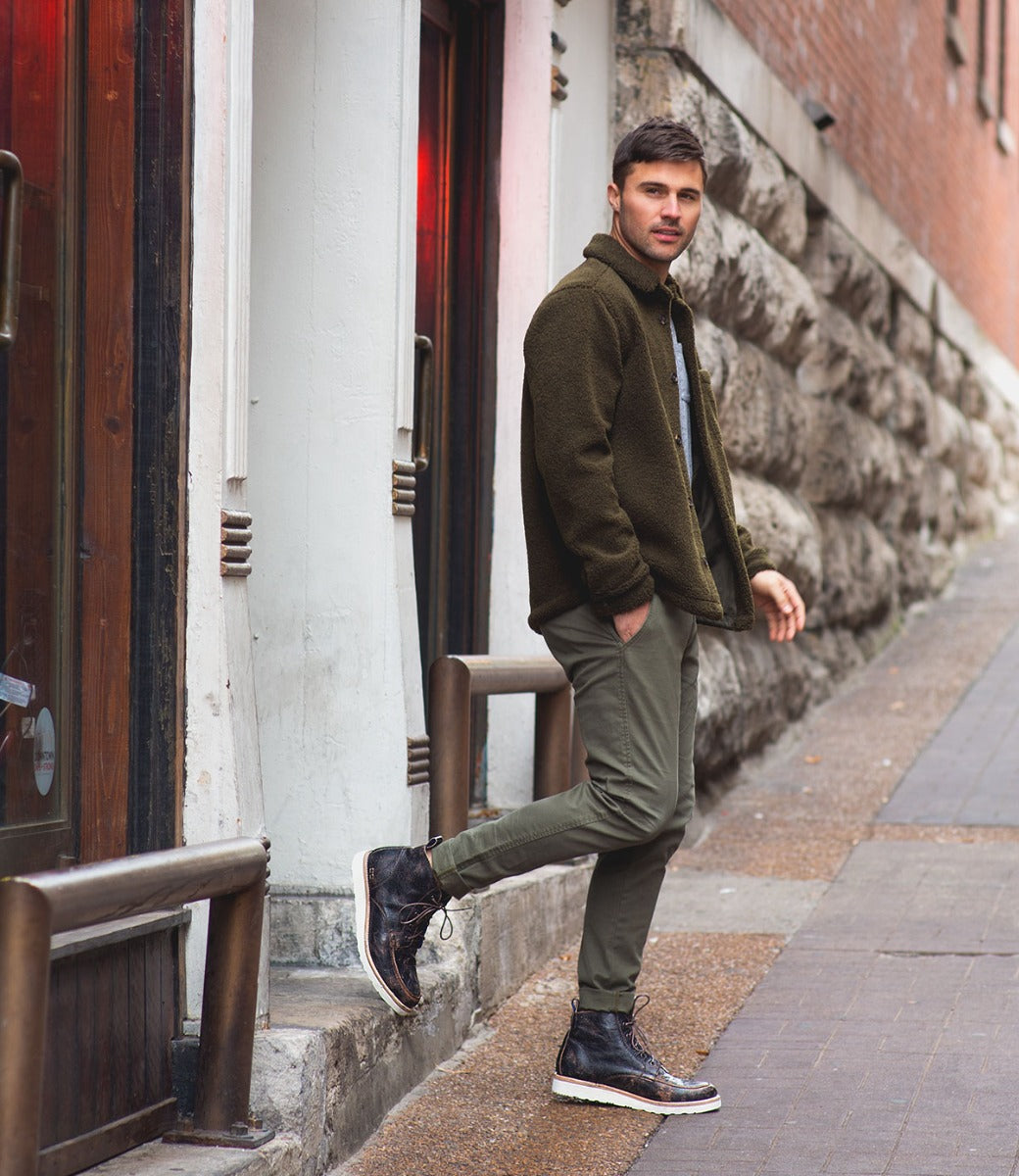 A man in a green jacket and pants leans against a brick wall on a city street, wearing Bed Stu Lincoln men's leather boots, and looking casually towards the camera.