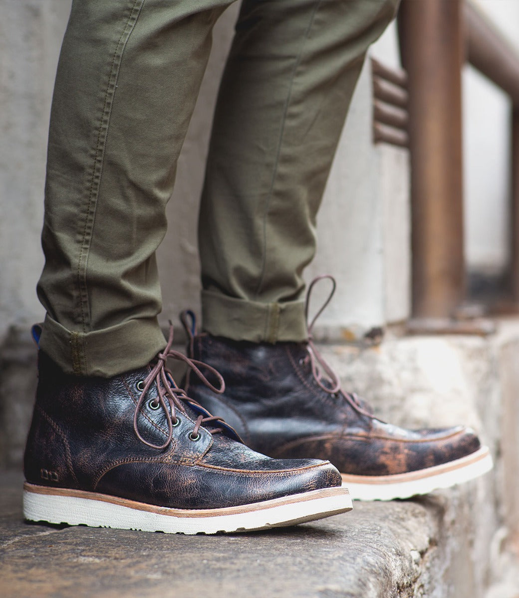 Close up of a person wearing Bed Stu Lincoln distressed brown men's leather boots and olive green pants, standing on concrete steps.