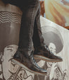 A person sitting on a ledge, wearing black jeans and Bed Stu men's leather boots, with a mural of a face in the background.