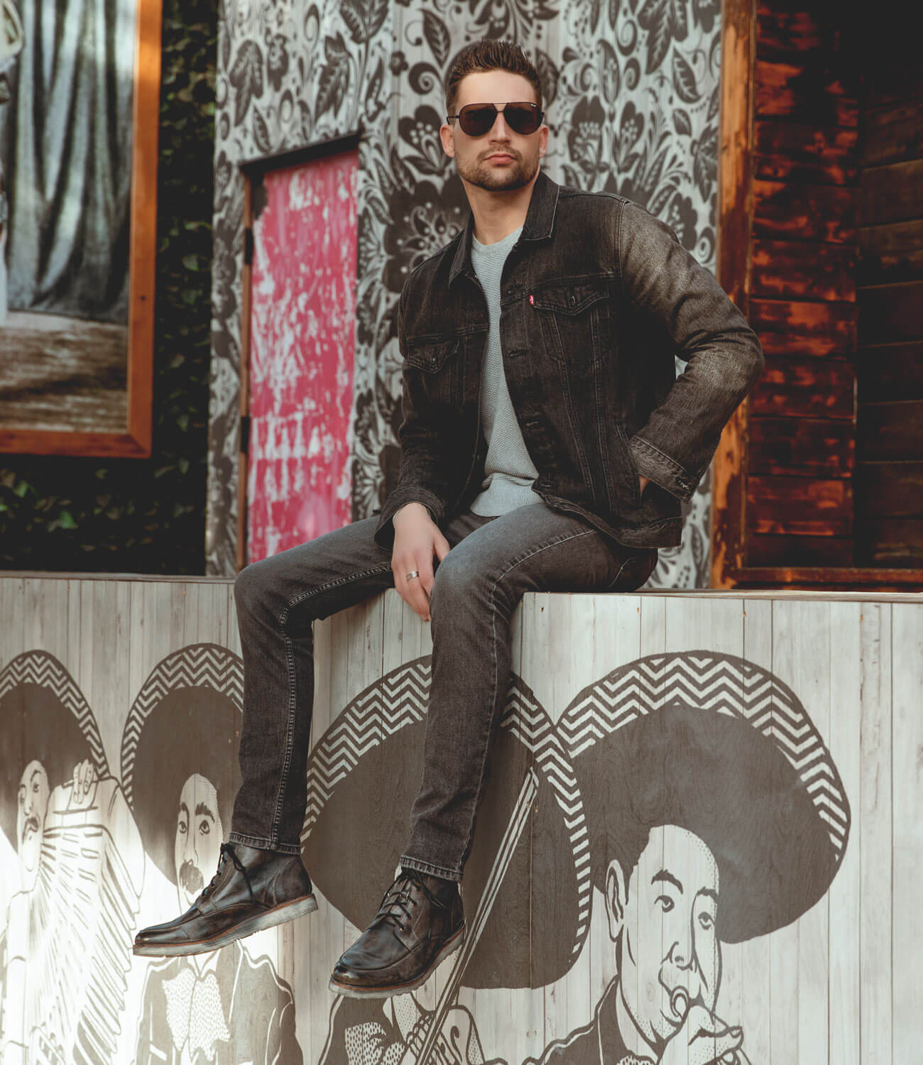 Man in sunglasses, denim jacket, and Bed Stu Lincoln work boots sitting on a wooden ledge in front of a wall with illustrated sombrero-wearing figures.