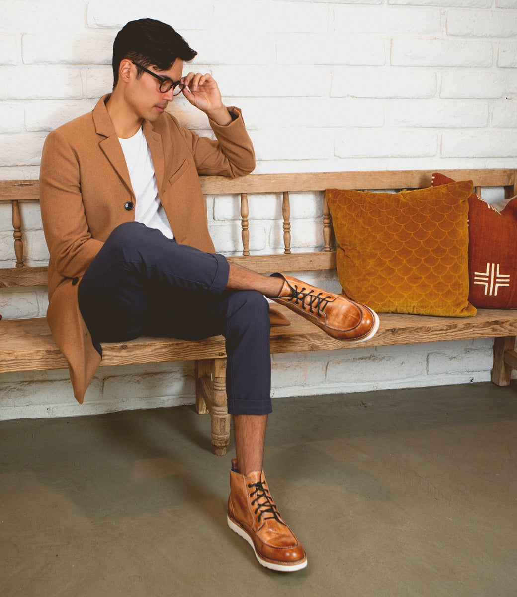 Man in a tan jacket and blue pants adjusting his glasses while sitting on a wooden bench with decorative pillows, wearing Bed Stu Lincoln men's leather boots.