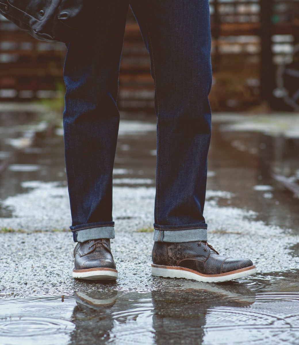 Person standing in a puddle wearing jeans and Bed Stu Protege Light Black Lux Boots with cushioned leather insoles, with wet ground and partial view of a wooden fence in the background.