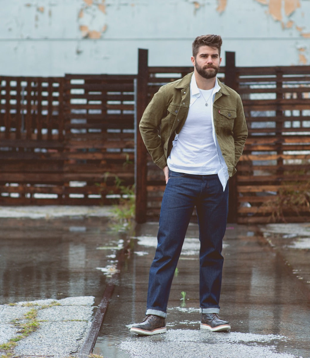 Man in a green jacket and jeans stands confidently in an urban setting with wet pavement and a rusted fence in the background, sporting Bed Stu Protege Light men's boots.