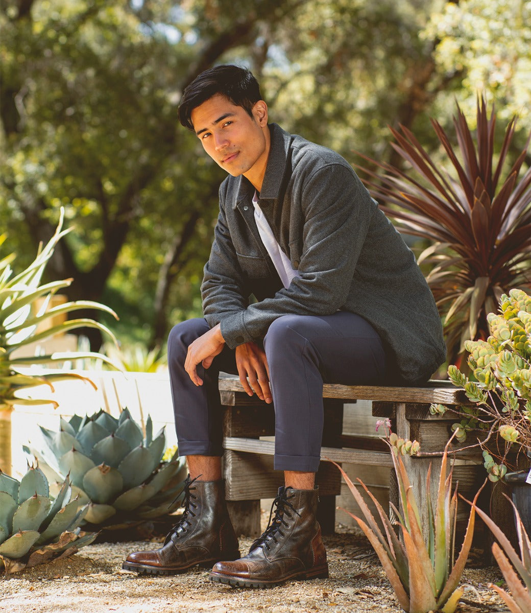 A young man sits on a wooden bench outdoors, dressed in a grey jacket and blue trousers, sporting Bed Stu Protege Trek combat style boots, surrounded by lush greenery.