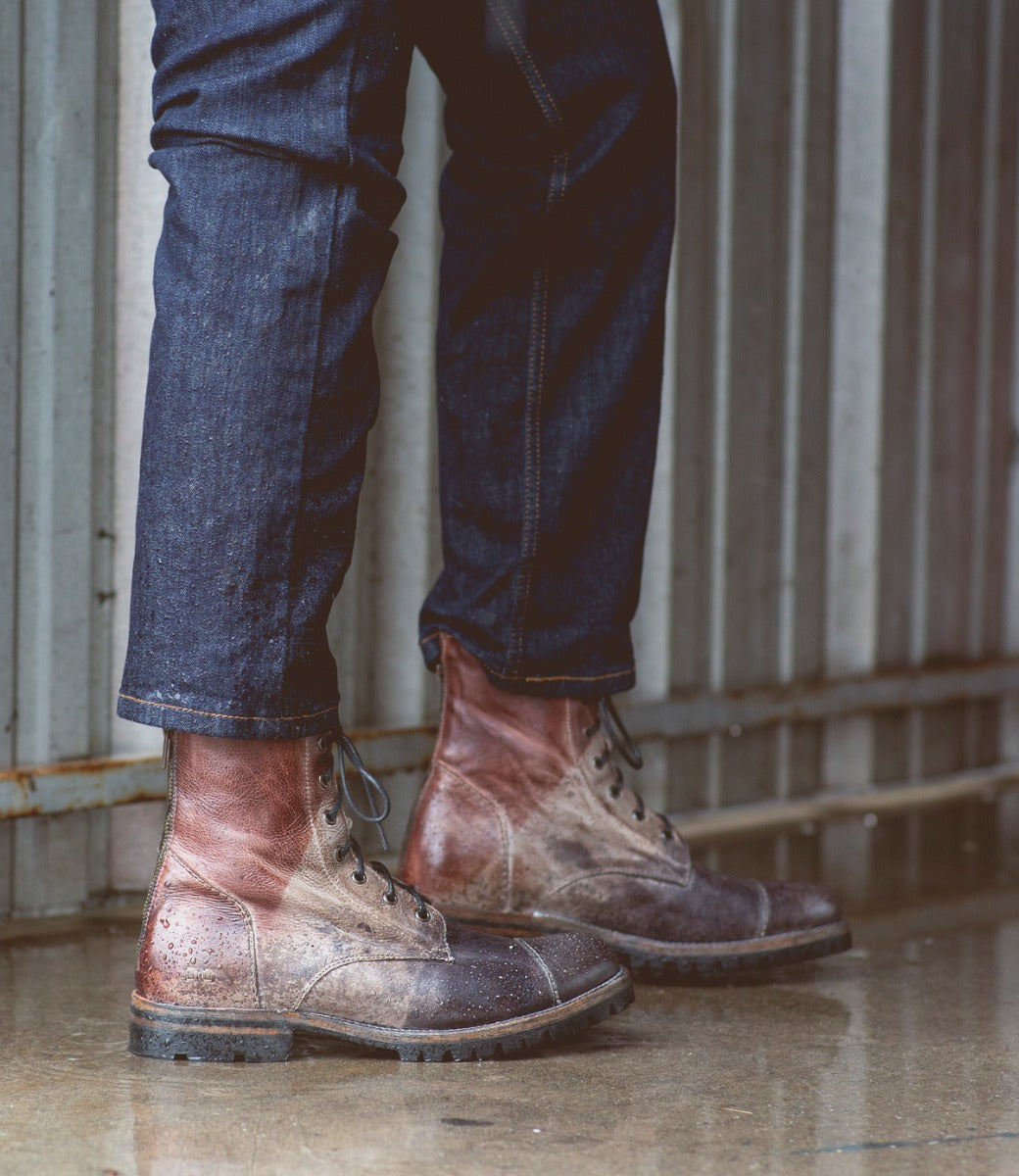 A close-up of a person's lower legs and feet wearing Bed Stu Protege Trek Teak Black Rustic Rust BFS boots with a Vibram outsole and blue jeans, standing on a wet surface.