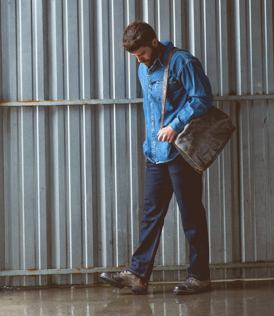 A man in a denim jacket and jeans carries a Bed Stu Protege Trek bag while walking by a corrugated metal wall in the rain, his combat style boots splashing through puddles.