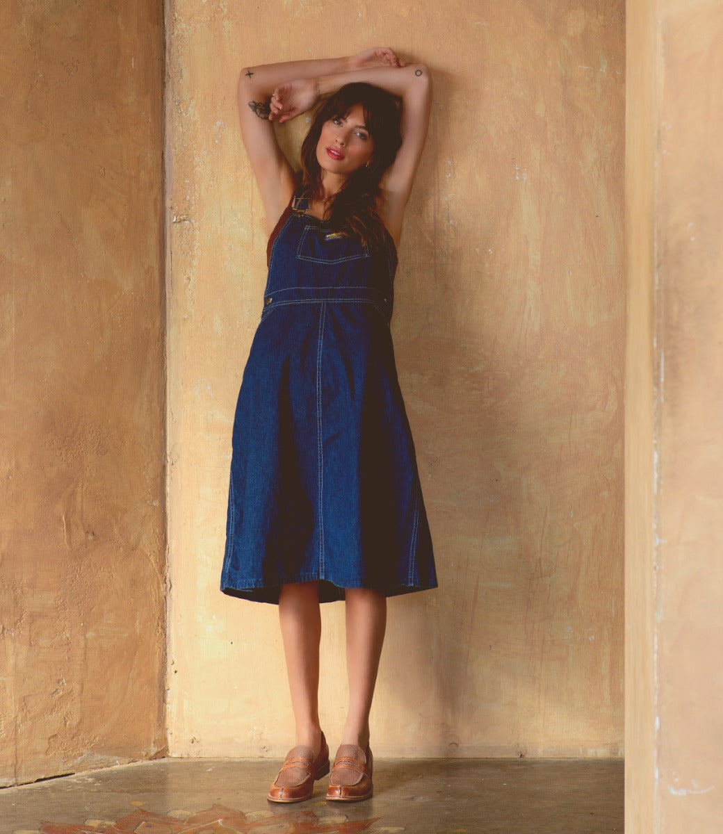 A woman in a denim overall dress leaning against a wall wearing Reina shoes by Bed Stu.