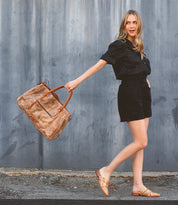 A woman in black shorts and sandals holding a Bed Stu Rockaway bag.
