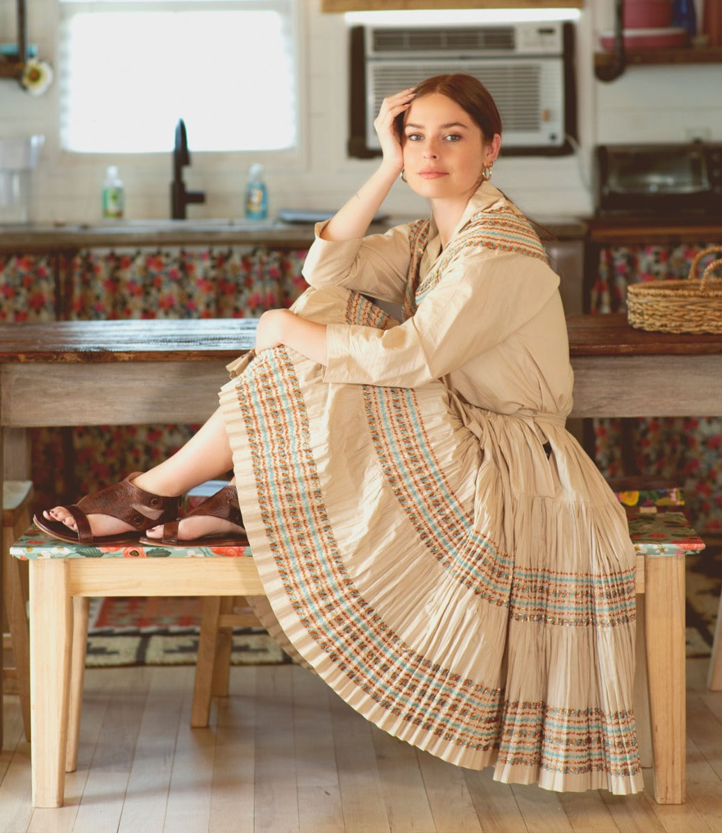 A woman in a tan dress sitting on a bench in a kitchen wearing Soto sandals by Bed Stu.