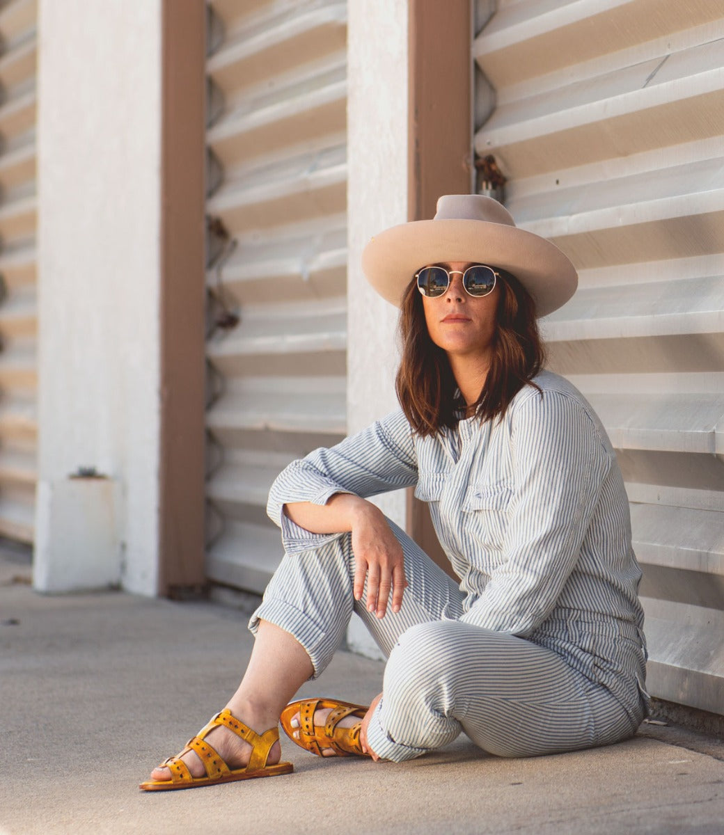 A woman sitting on the sidewalk wearing a Sue hat and Bed Stu sunglasses.