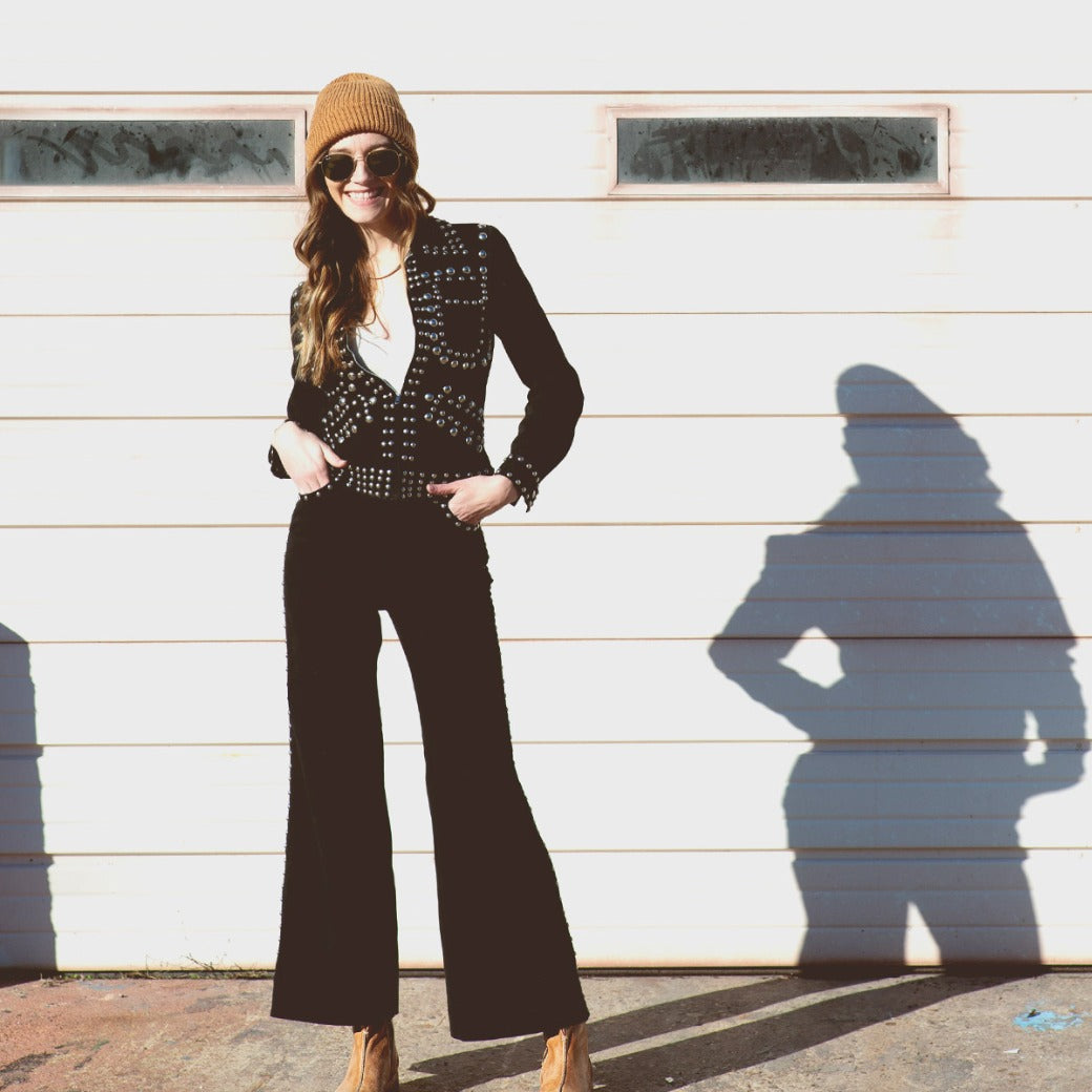 A woman is standing in front of a garage wearing black pants and a beanie from Bed Stu.
