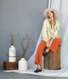 A woman wearing a Tania hat and orange pants sits on top of a Bed Stu wooden box.