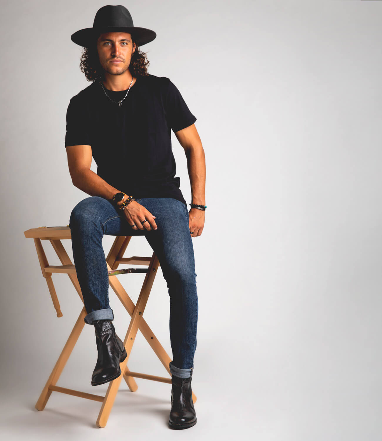 A man in a black outfit and wide-brimmed hat sits on a wooden stool against a gray background, wearing Bed Stu leather ankle boots, looking at the camera.
