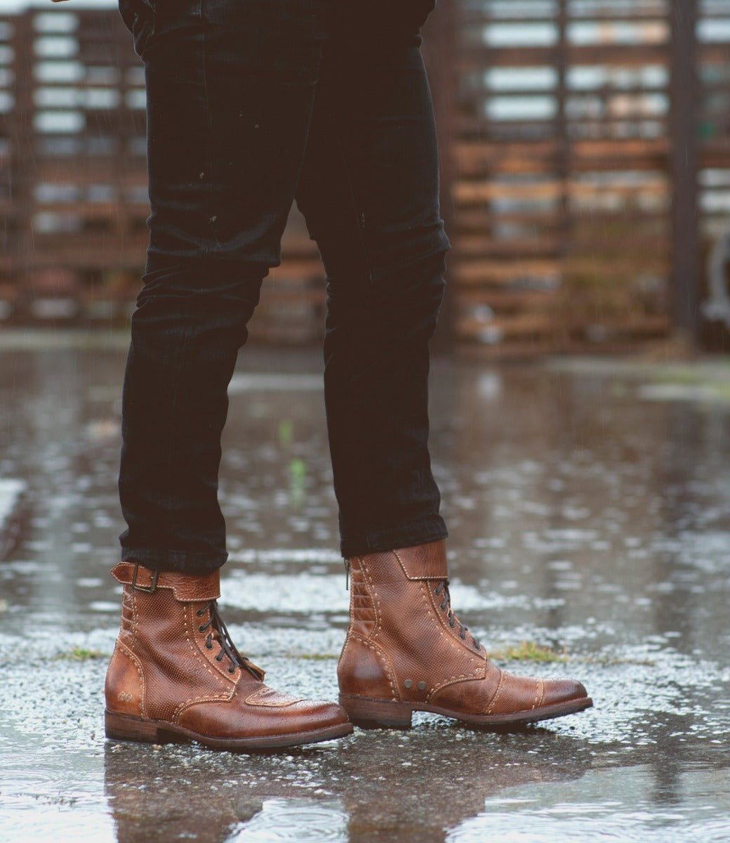 A person wearing Bed Stu's brown leather moto-inspired boots, "Walker," and black jeans standing on a wet surface with puddles, reflecting a cloudy sky.