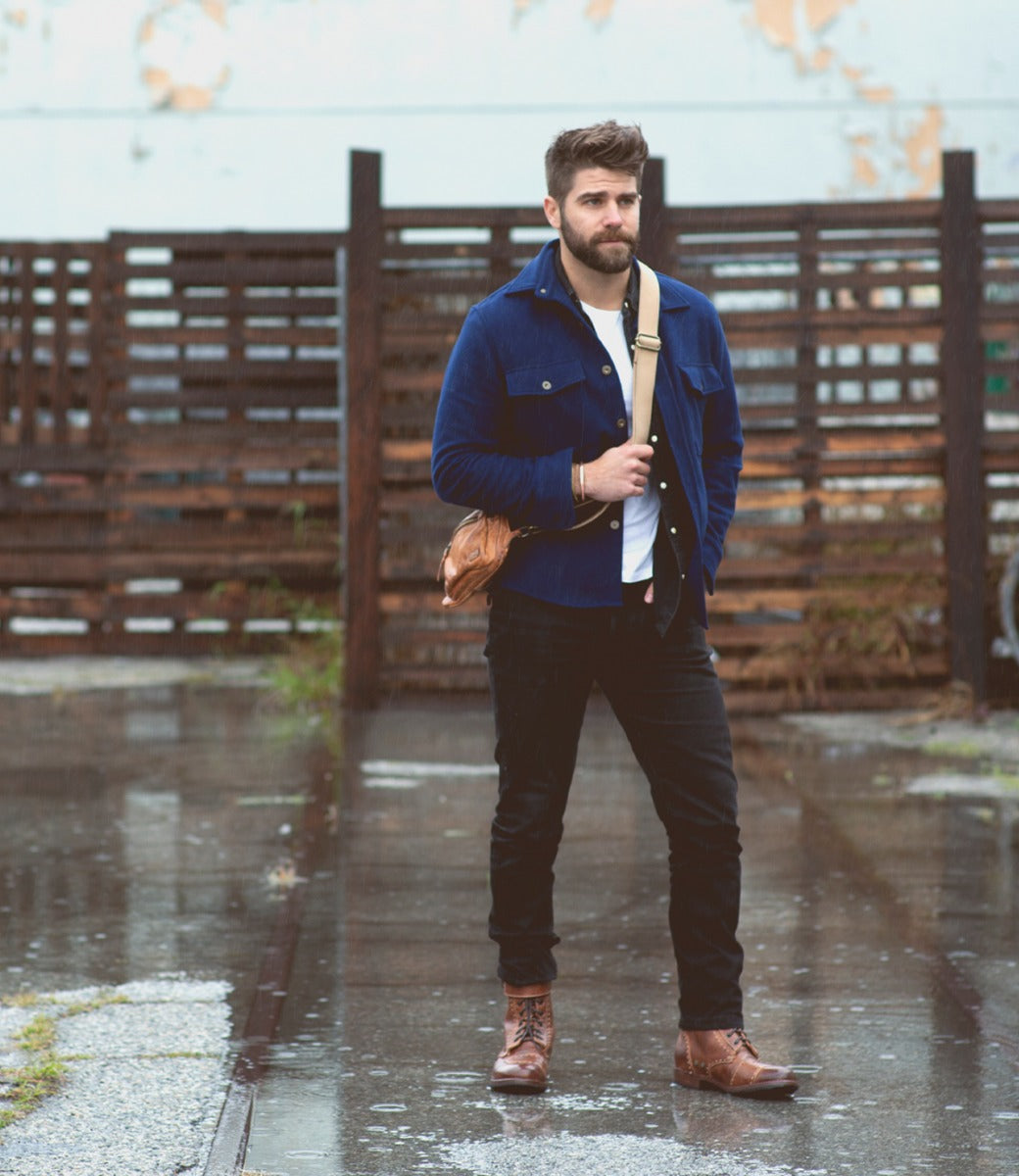 A man in a blue jacket, dark jeans, and moto-inspired boots with VIBRAM® lug outsole standing on a wet street, holding a leather bag, with a rustic fence in the background.