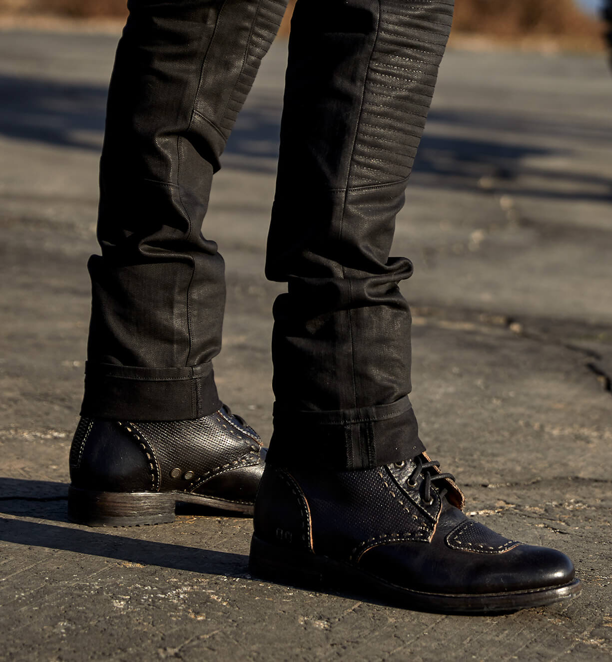 Close-up of a person wearing Bed Stu moto-inspired boots and black jeans standing on a cracked pavement.
