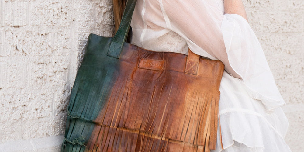 Person in a flowing white dress carries a large brown leather tote with dark green side panels and a textured, fringed design.