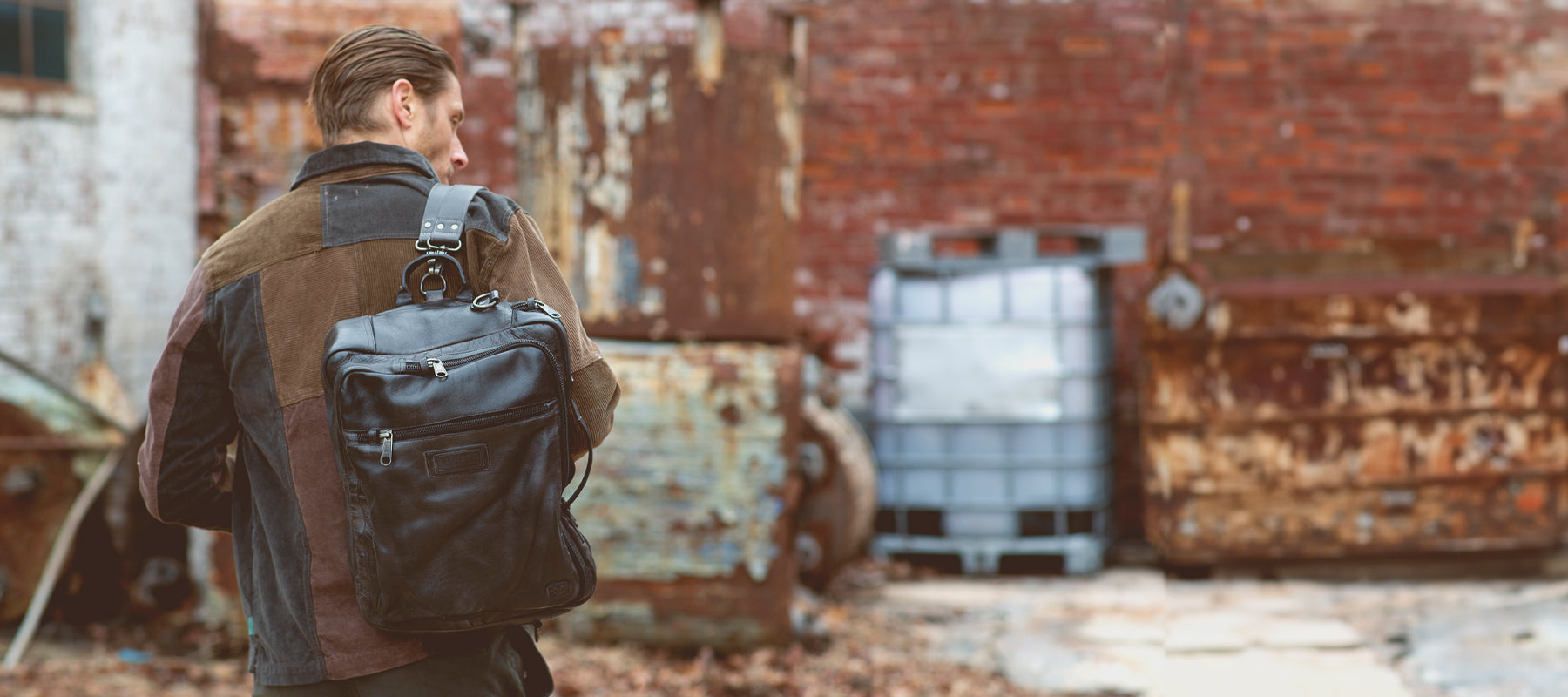 A brunette man in a patchwork leather jacket carrying a black Bed|stu leather backpack in a rustic brick alley.