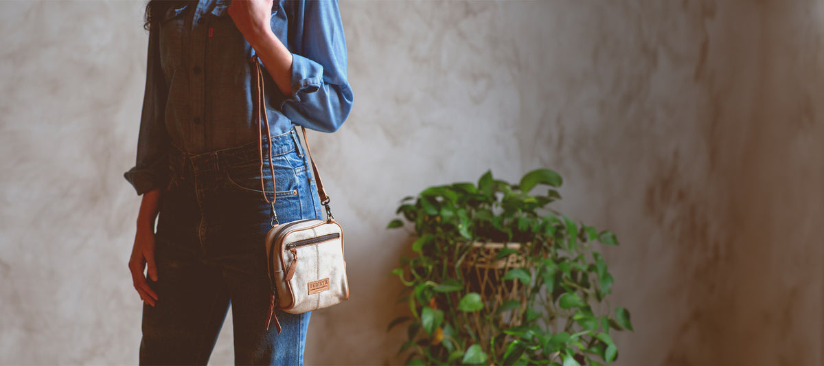 A brunette woman in a blue jean vest and white skirt with tan bag across her body in lush landscape.