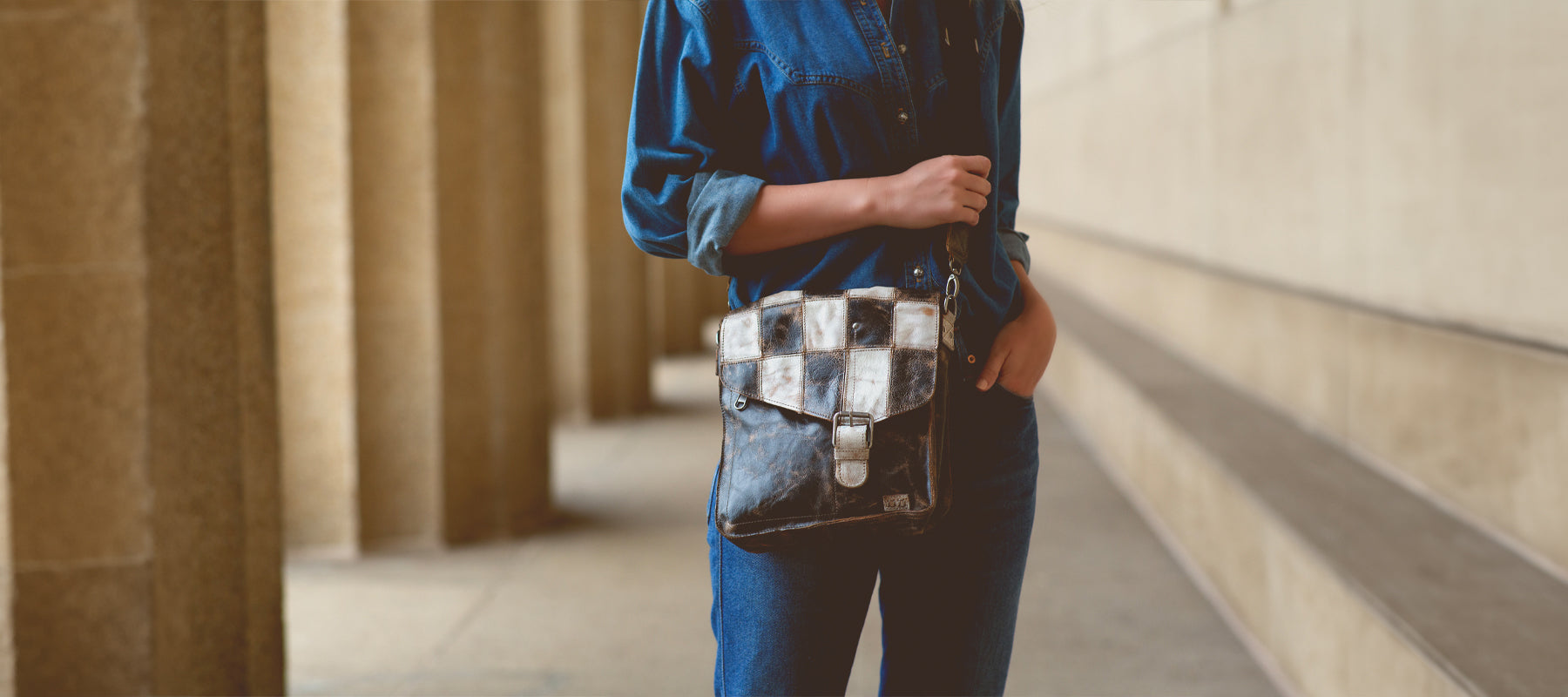 A brunette woman standing by a rock outcropping, wearing sunglasses, blue vest, white skirt, and blue leather bag.
