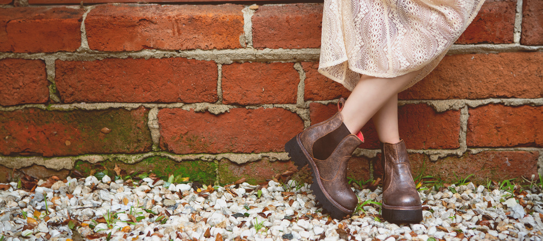 Person leaning against a brick wall wearing brown boots and a lace dress.
