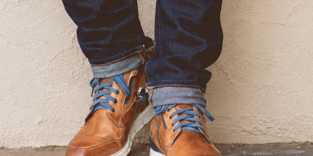 Person wearing brown leather lace-up ankle boots with blue laces, white soles, and dark cuffed jeans.