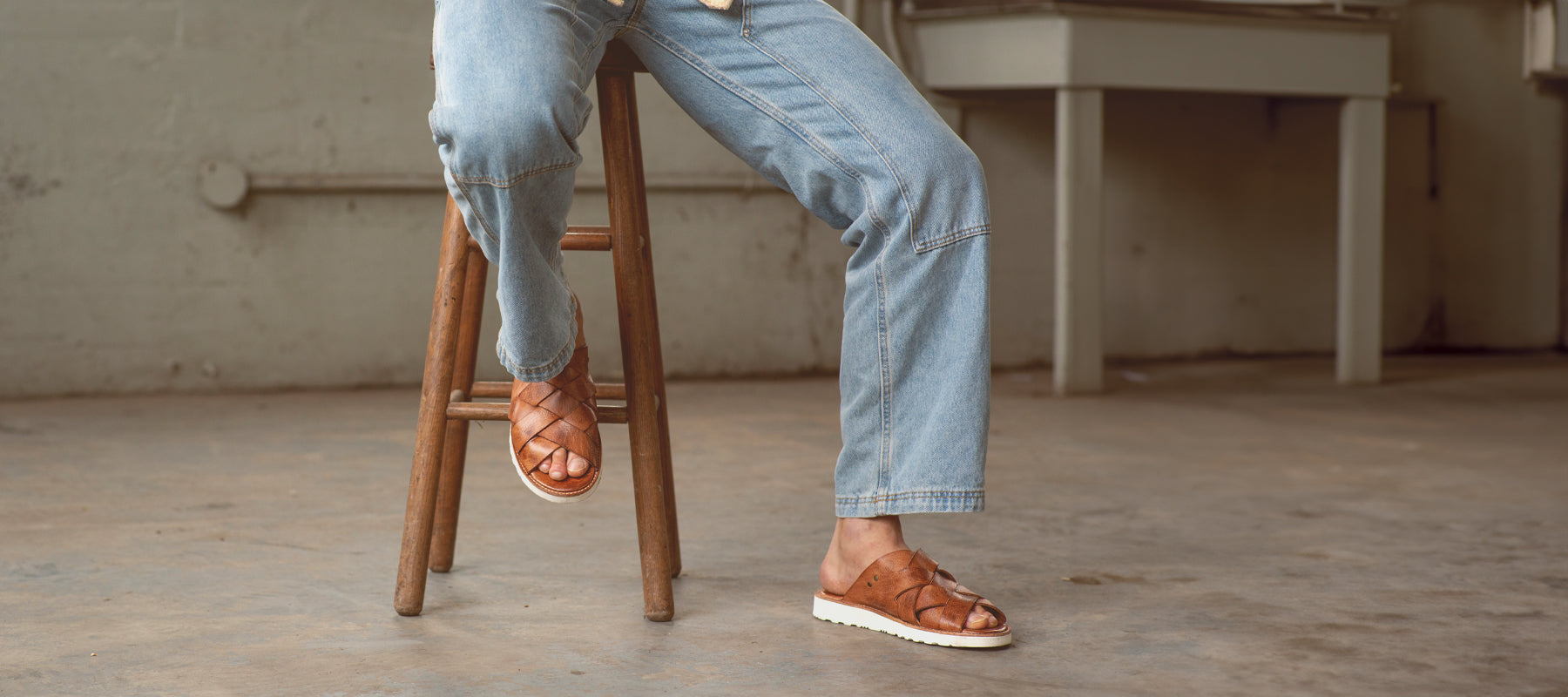 A brown haired man sitting at a small coffee table on a front porch while wearing a black shirt, blue jeans, and black distressed sandals.