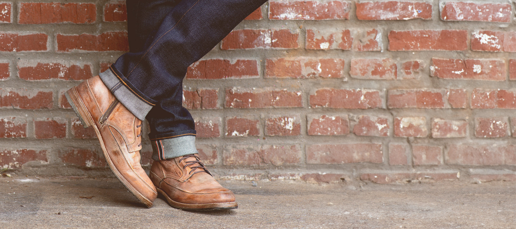 A man in dark denim heans with a cuff in a tan lace-up boot standing in front of a brick wall