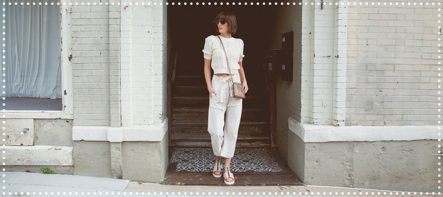 Woman in white crop top and trousers standing in a shadowed doorway, holding a beige purse, urban background.