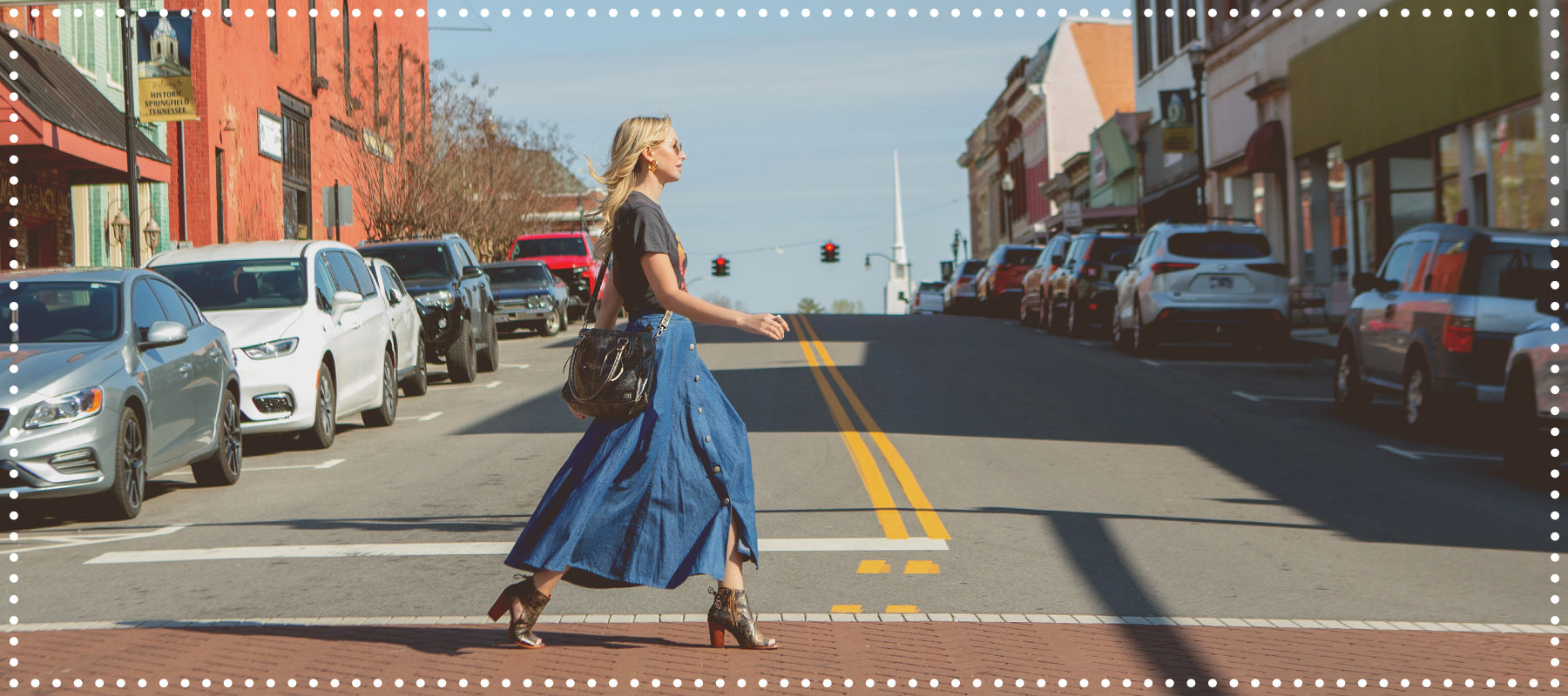 A woman crossing a street in a small town, wearing a denim skirt and carrying a backpack, with cars parked along the roadside.