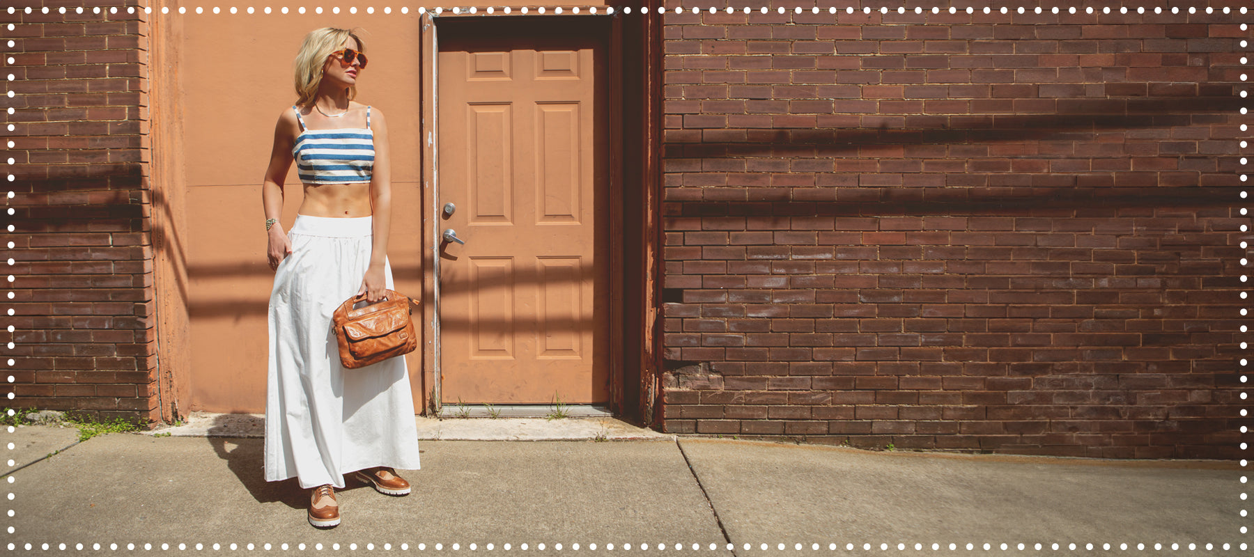 Woman in a striped top and white pants standing beside a red brick wall and a brown door, holding a brown bag and wearing sunglasses.
