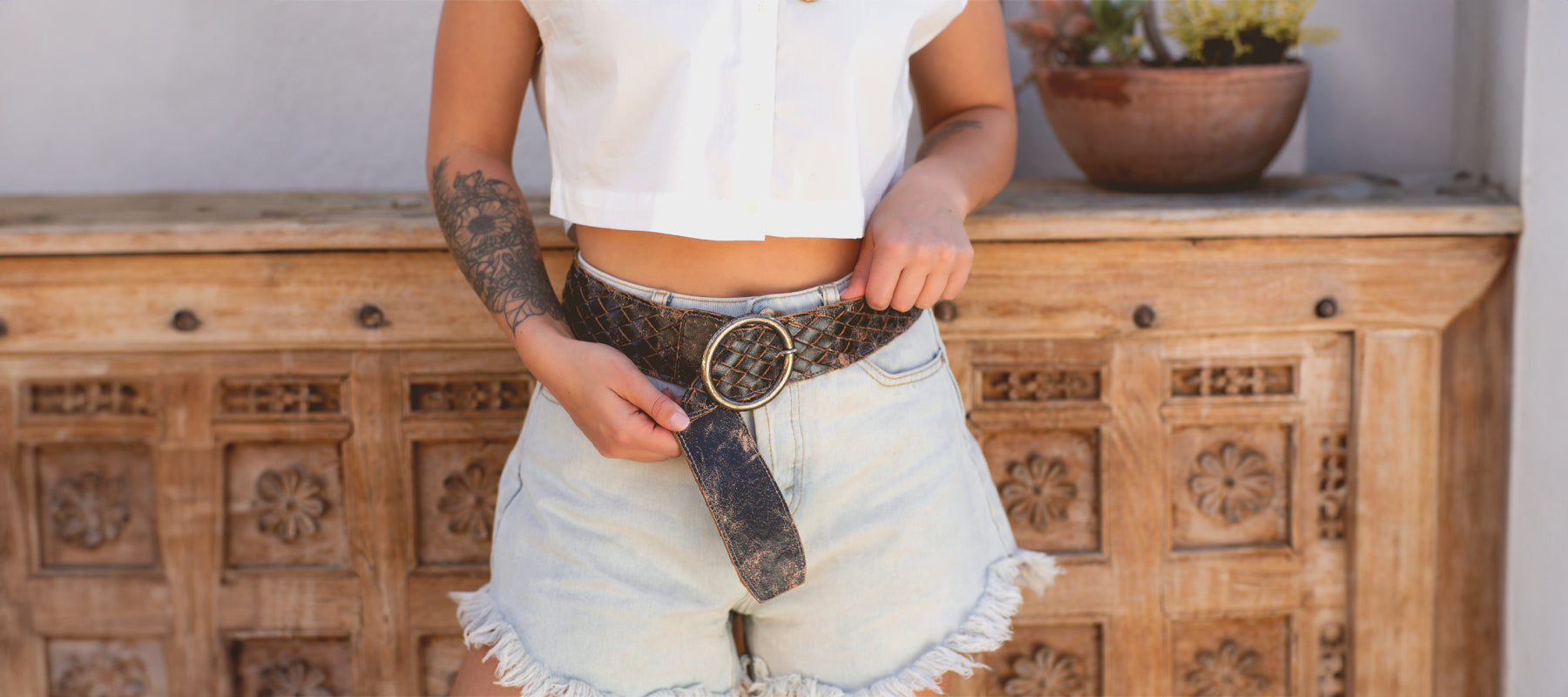 A person in a white crop top and denim shorts, showcasing a tattooed arm and holding a stylish belt, standing in front of a carved wooden cabinet.