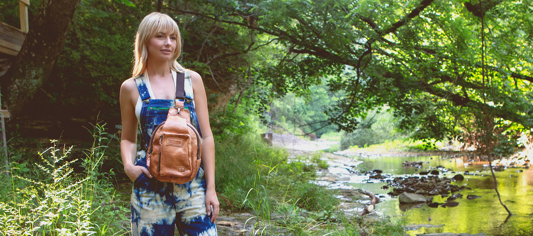 Blonde woman standing by a creek while wearing blue and white tie dye overalls and tan leather backpack.