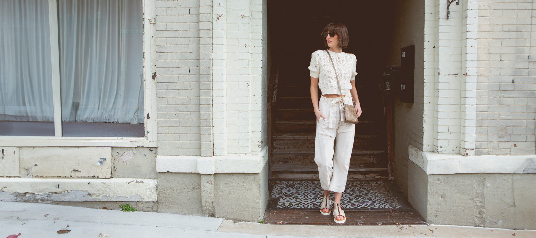 A knee-down picture of a woman with a knee'-length white skirt on a concrete floor iwth distressed blue leather sandals on next to a large ceramic pot. 