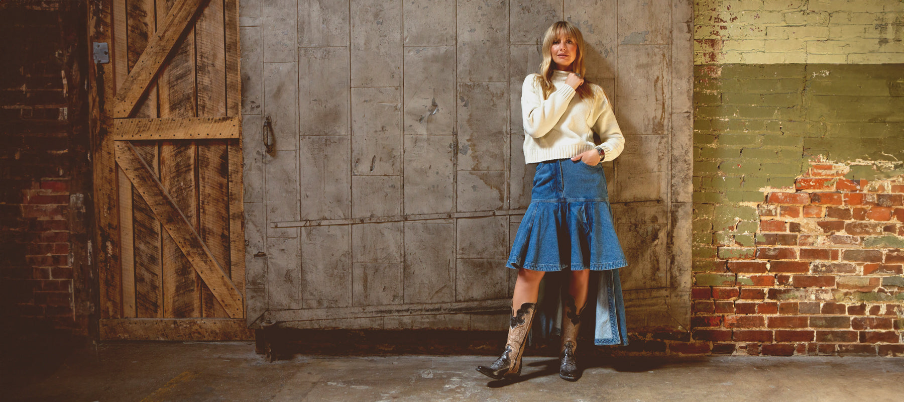 A blonde woman standing in front of a rustic wood and brick wall in a knee-length jean skirt, white sweater and Queen Bedstu western boots. and brick