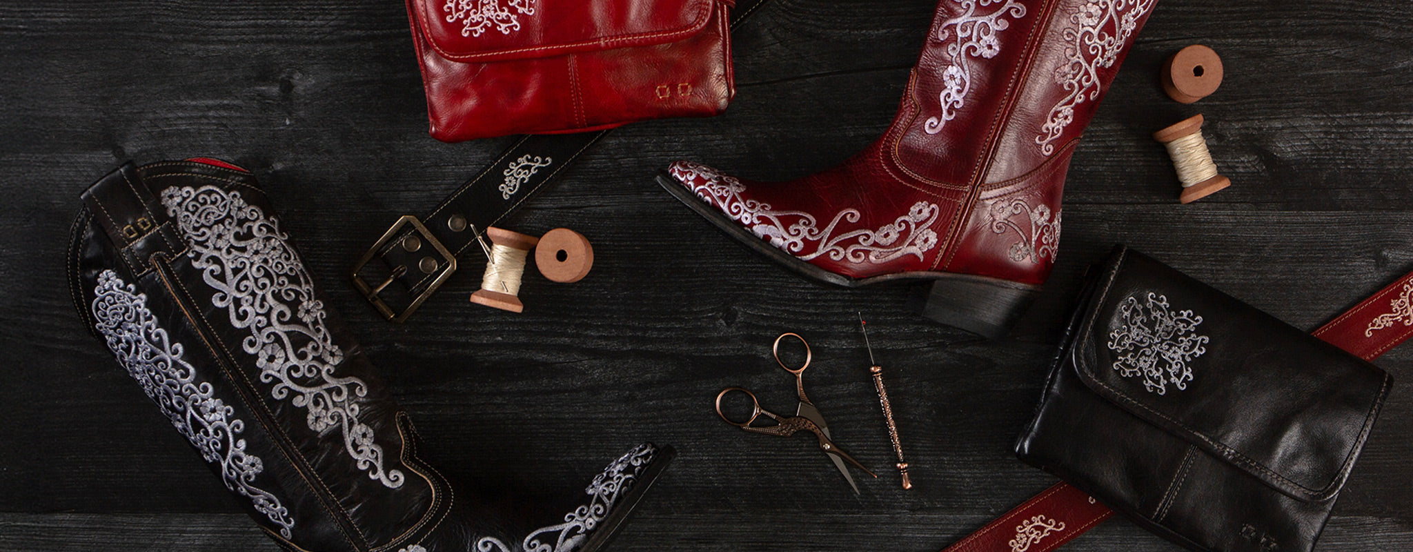 Red and black embroidered leather boots, matching belts, wallets, spools of thread, and scissors arranged on a dark wooden surface.