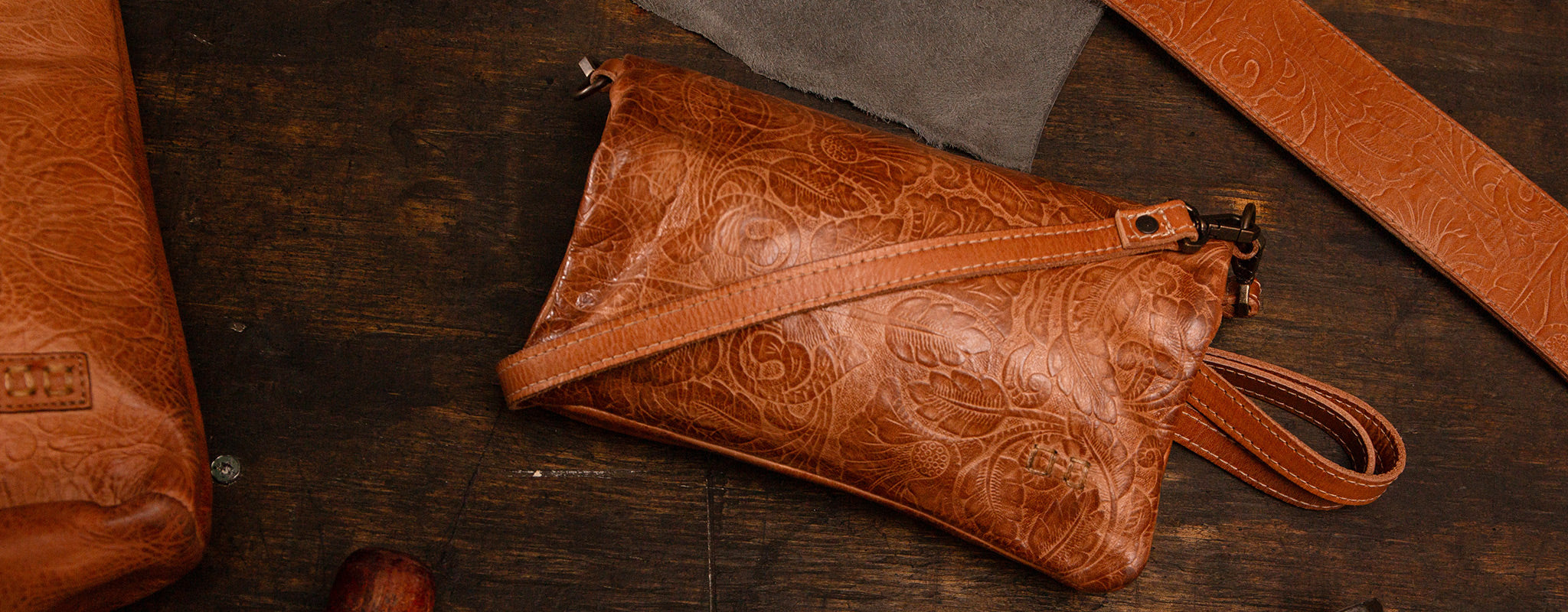 A brown leather wristlet with embossed floral patterns lies on a wooden surface alongside scraps of leather and tools.