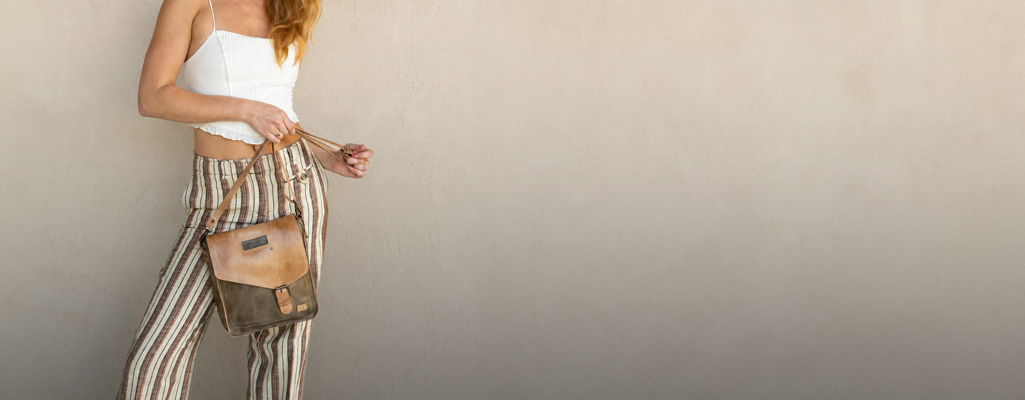 Person wearing a tan-and-olive leather crossbody bag with striped pants and a white top, standing against a neutral beige wall.