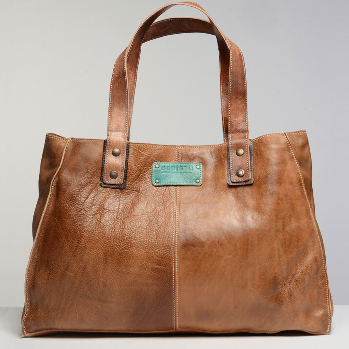 The Studio Arlington Body, a brown leather tote bag by Bed Stu, features two handles and a stitched rectangular patch with the text "BEDSTU." Rivet details at the handle bases add an extra touch of style. Displayed against a plain background, this personalized bag is perfect for showcasing your own style and creativity.