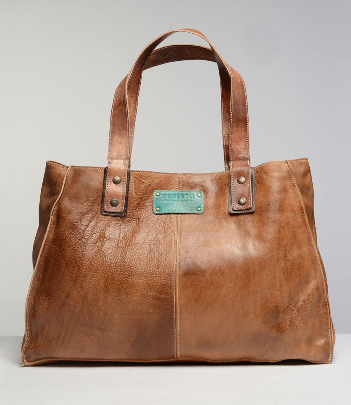 The Studio Arlington Body, a brown leather tote bag by Bed Stu, features two handles and a stitched rectangular patch with the text "BEDSTU." Rivet details at the handle bases add an extra touch of style. Displayed against a plain background, this personalized bag is perfect for showcasing your own style and creativity.