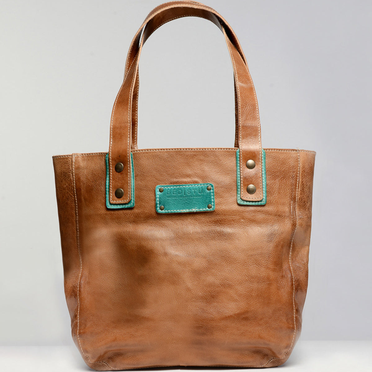 The Studio Gaviota Body by Bed Stu is a tan leather tote bag featuring two handles and a rectangular label with teal accents on the front. Customize your own personalized bag by choosing from a variety of colors, making it uniquely yours.