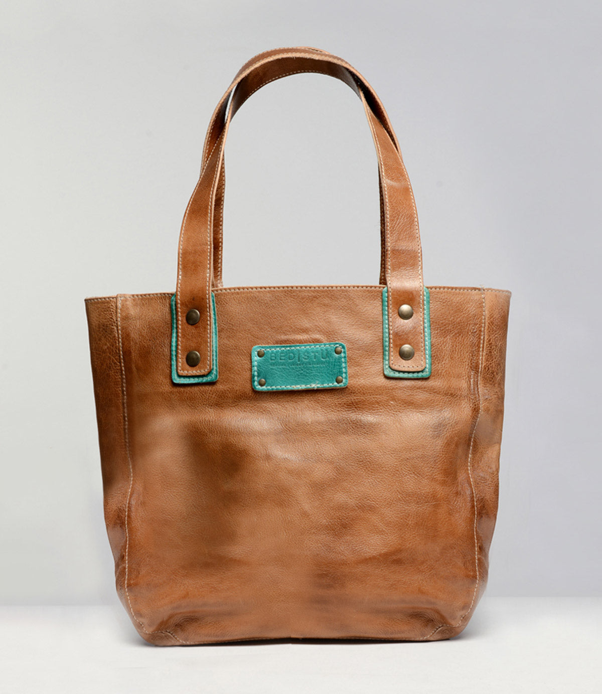 The Studio Gaviota Body by Bed Stu is a tan leather tote bag featuring two handles and a rectangular label with teal accents on the front. Customize your own personalized bag by choosing from a variety of colors, making it uniquely yours.