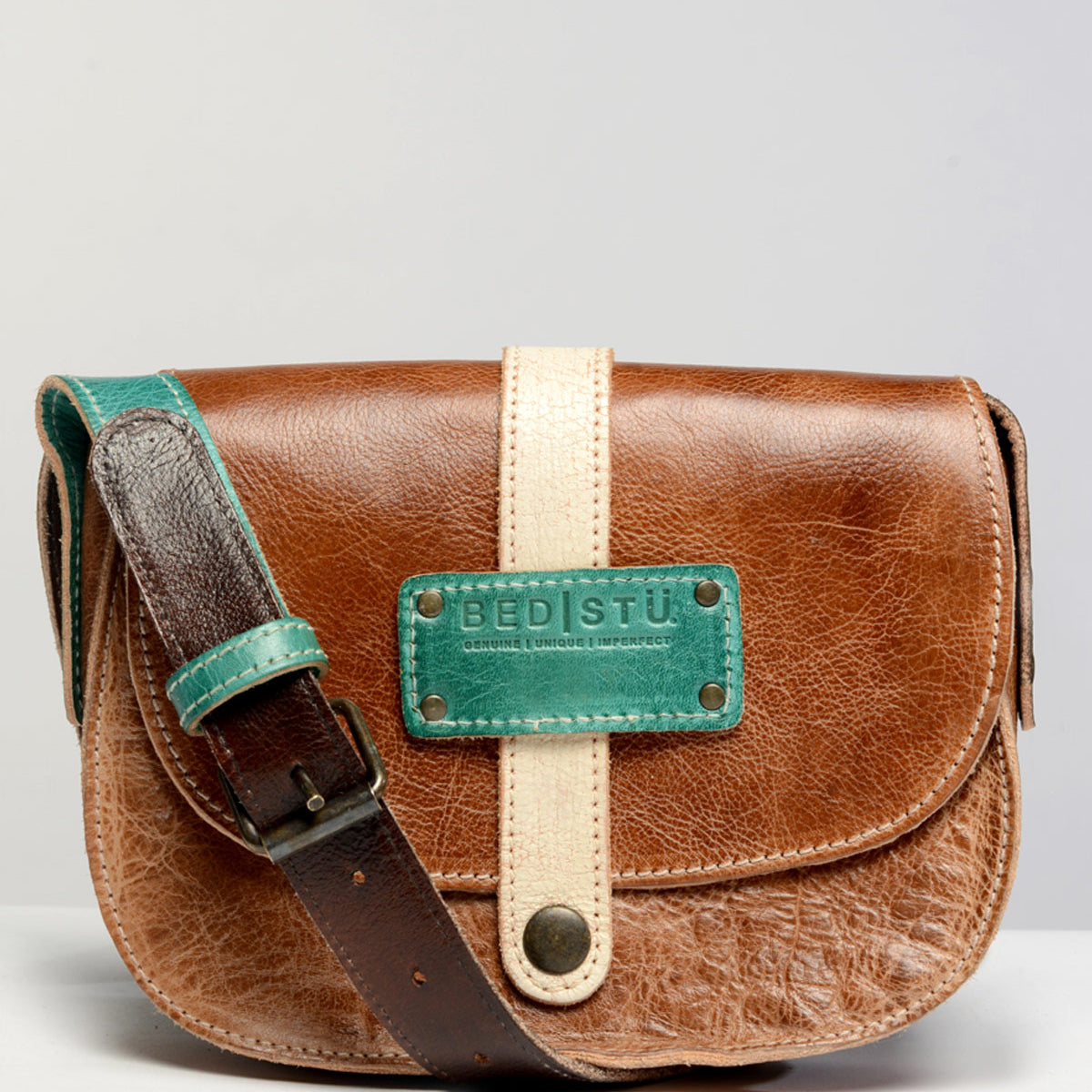 The Studio Shoreline Body, by Bed Stu, is a brown leather crossbody bag with a green and beige front strap, featuring a metal clasp and a green brand label reading "Bed Stu." This personalized bag includes a brown strap with a green adjustment section.