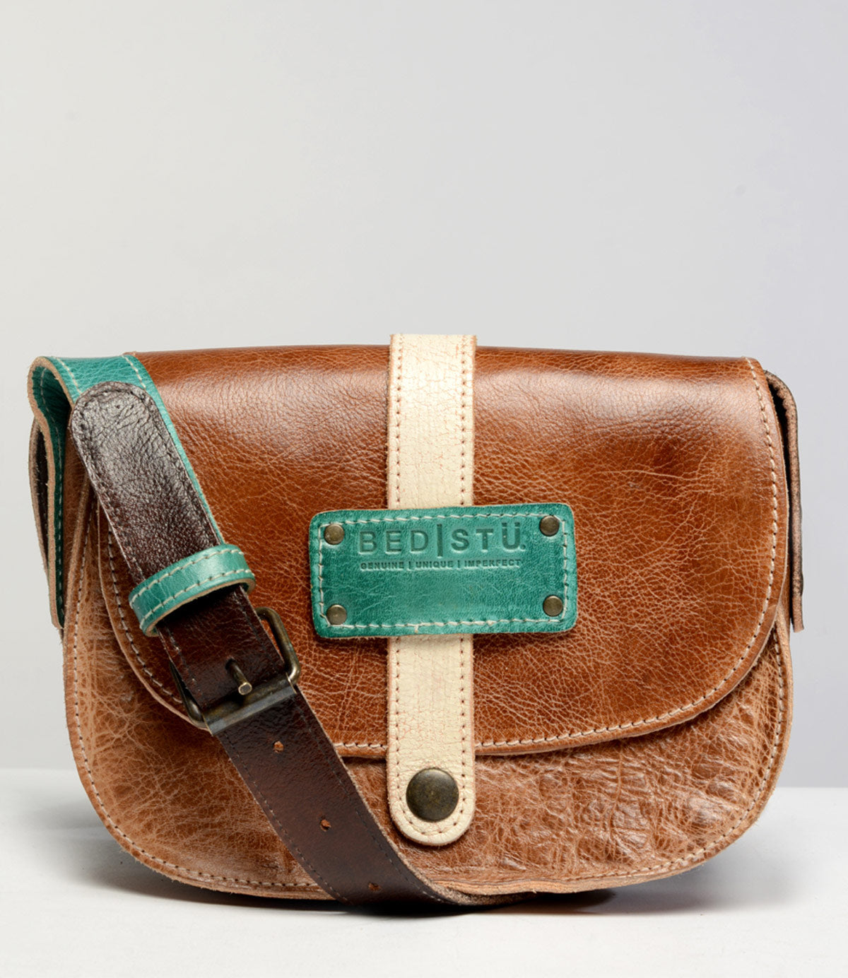 The Studio Shoreline Body, by Bed Stu, is a brown leather crossbody bag with a green and beige front strap, featuring a metal clasp and a green brand label reading "Bed Stu." This personalized bag includes a brown strap with a green adjustment section.