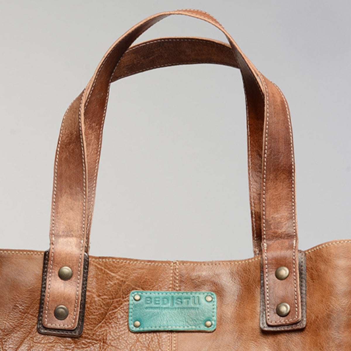 A close-up of a brown leather Studio Short Shoulder Strap bag with double handles and a light blue label reading "Bed Stu," perfect for those who value creativity in personalized bags.