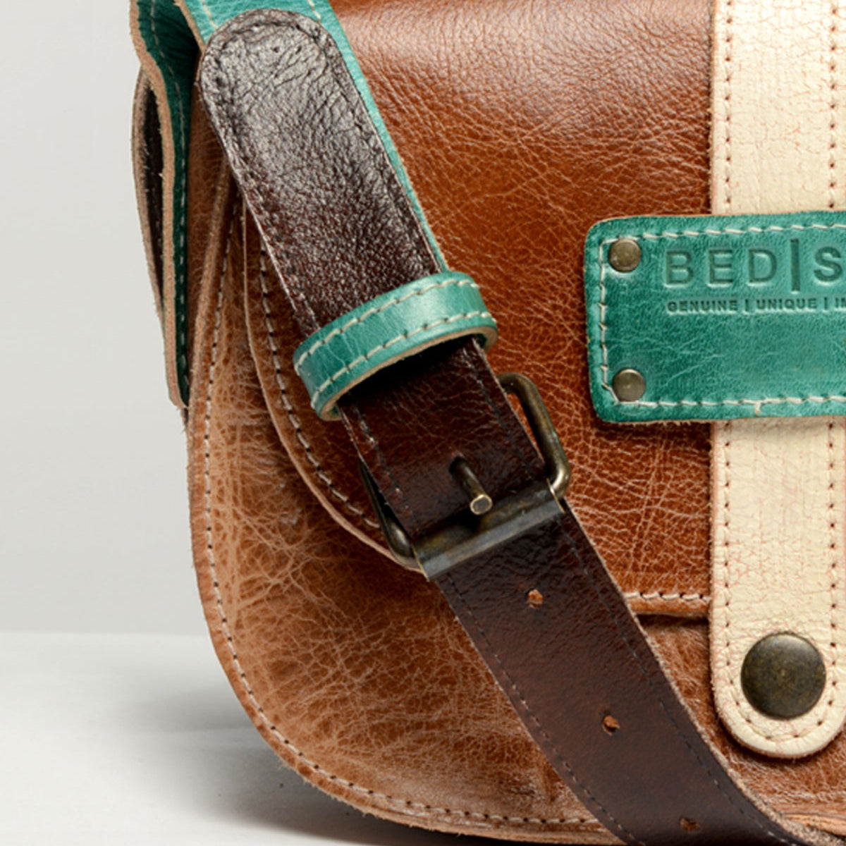 Close-up of a brown leather satchel with a turquoise strap and a beige vertical strap, showcasing textured details and a small metal buckle. A label with "Bed Stu" is partially visible, hinting at the creativity behind this personalized bag named "Studio Long Shoulder Strap.