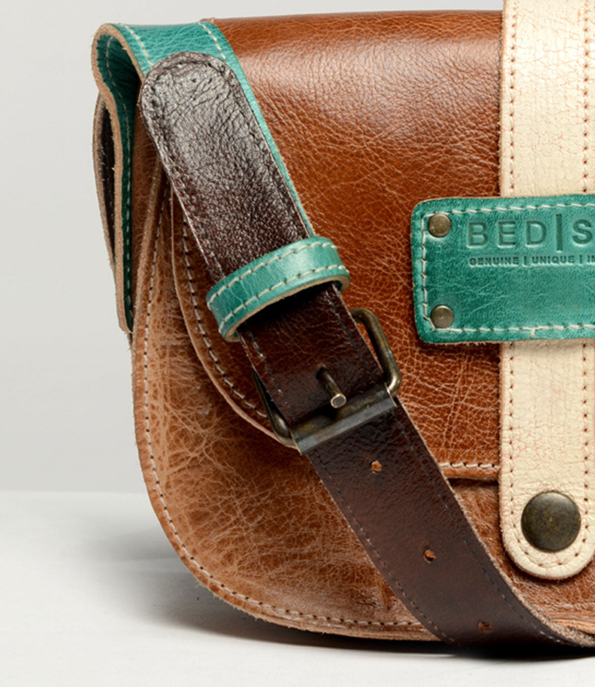 Close-up of a brown leather satchel with a turquoise strap and a beige vertical strap, showcasing textured details and a small metal buckle. A label with "Bed Stu" is partially visible, hinting at the creativity behind this personalized bag named "Studio Long Shoulder Strap.